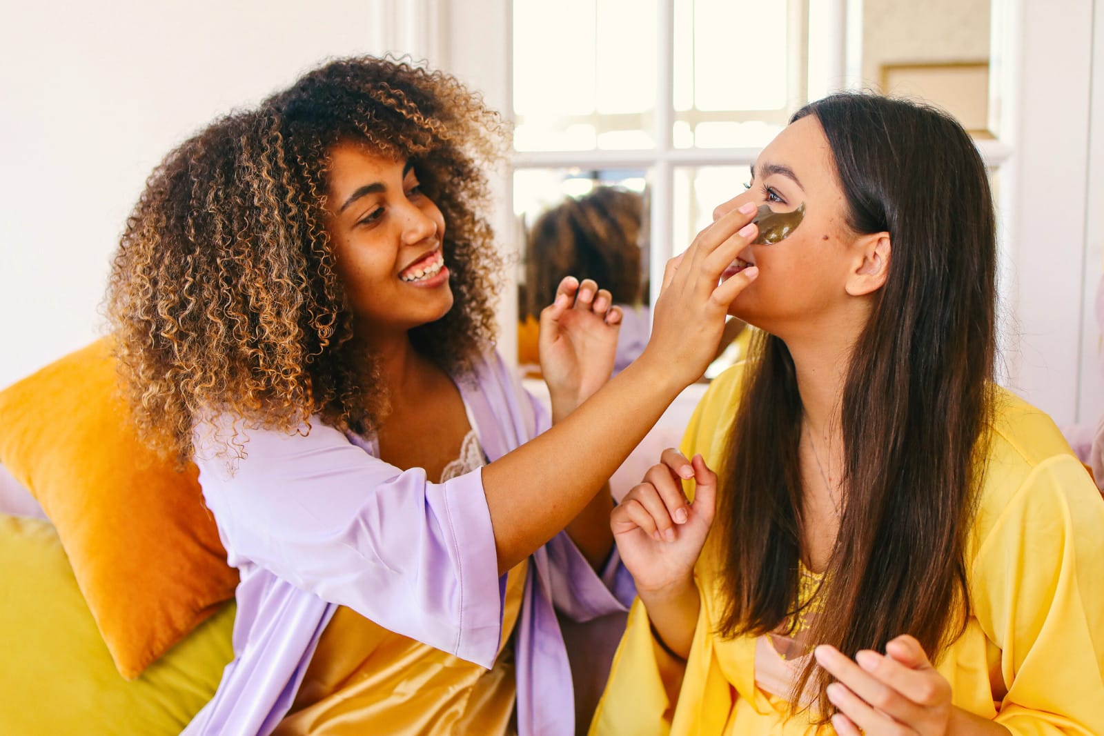 Two friends sitting on a yellow couch, one with curly hair in a purple shirt and the other with straight hair in a yellow shirt, have a self-care routine. Image @Canva