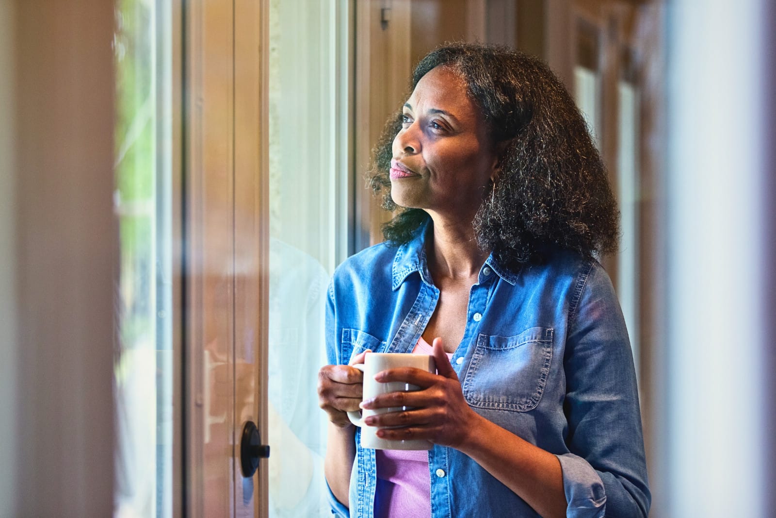 A woman standing near a glass door, holding a mug, and contemplating. Image @Canva