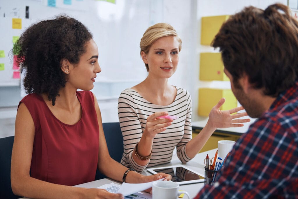 Three individuals engaged in a discussion around a table with colorful sticky notes in the background. Image @Canva