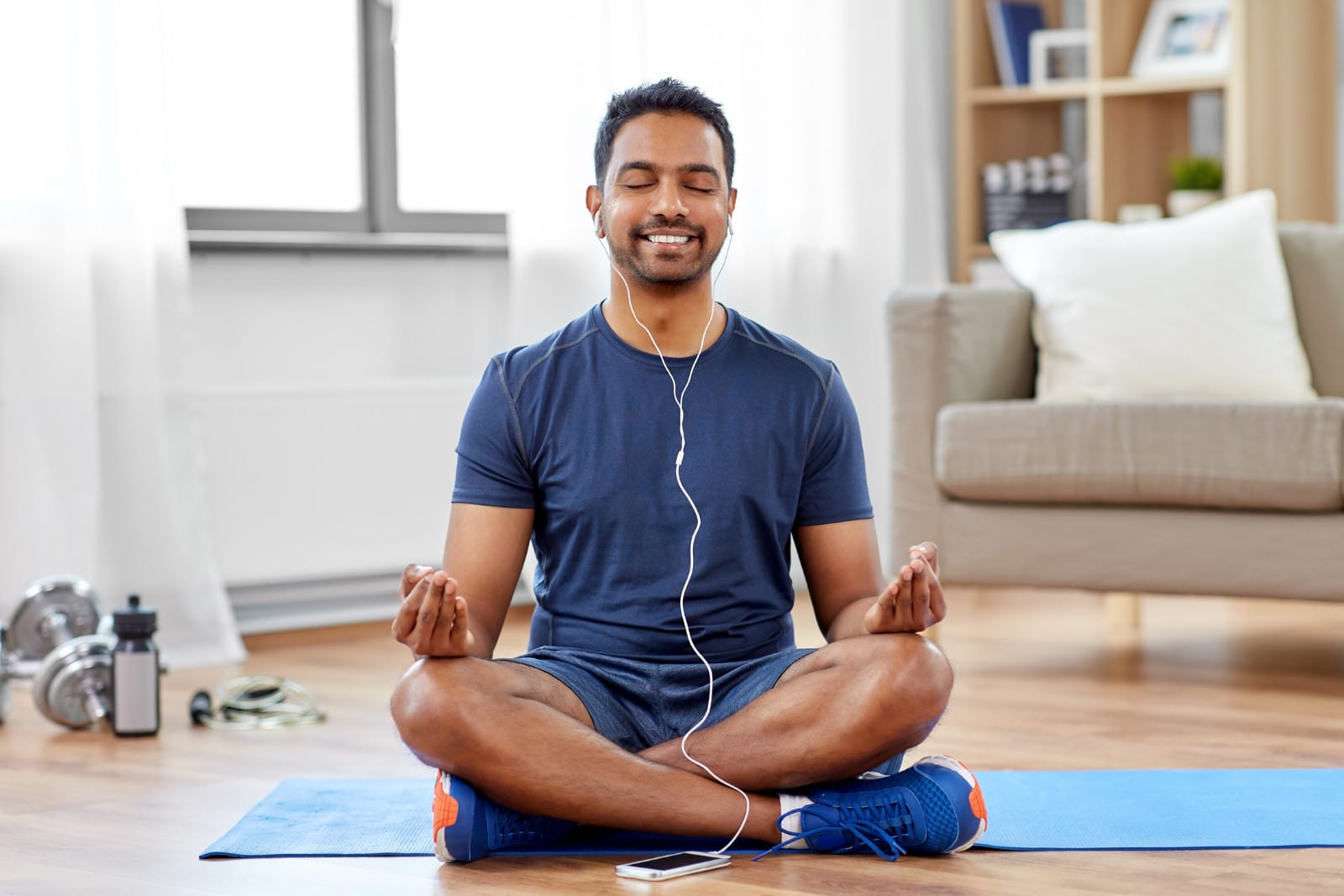 An man sitting cross-legged on a blue yoga mat, meditating or relaxing with earphones in a room with hardwood flooring. Image @Canva