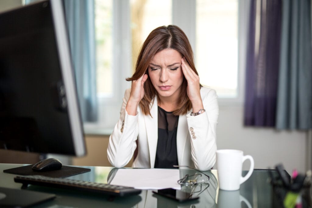 Person sitting at a desk with their head in their hands, trying to remain calm. A white mug and papers are on the desk, with a computer monitor to the side. Image @Canva