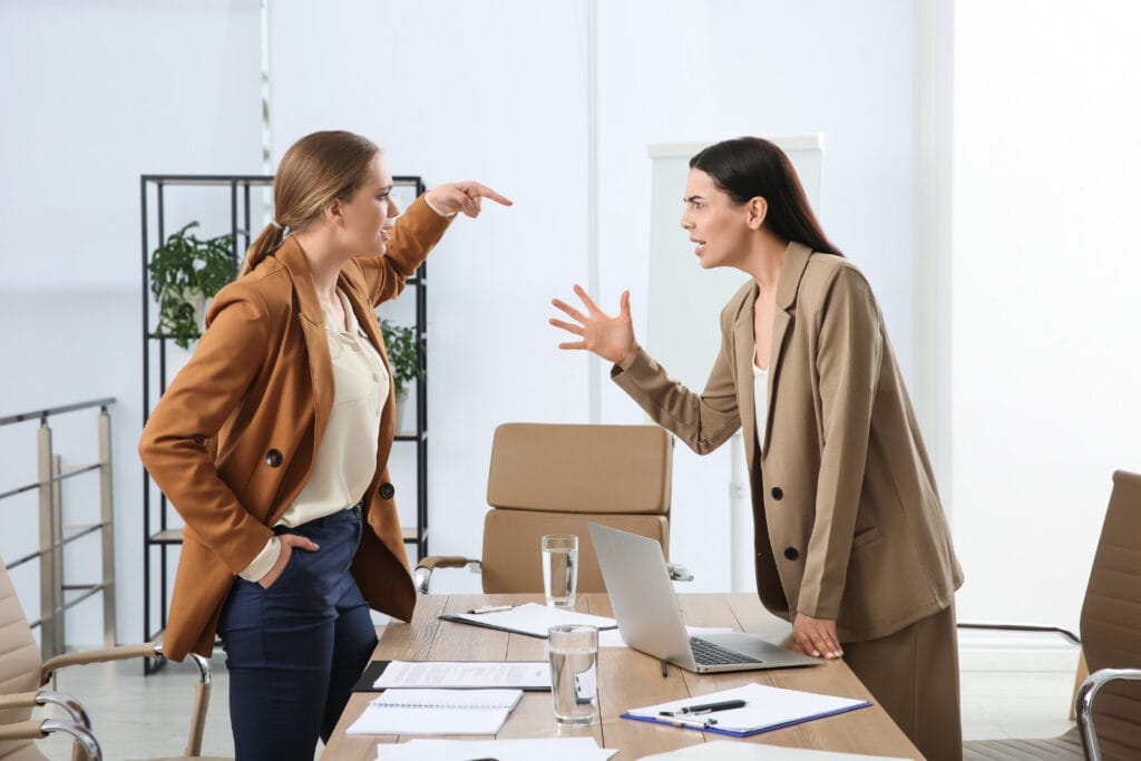 Two individuals in an office setting, one pointing a finger while the other responds with an open hand gesture. Image @Canva