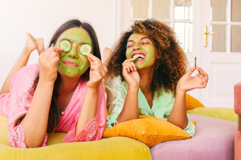 Two women enjoying a relaxing spa day at home. One applies cucumber slices to her eyes, while the other bites into a cucumber slice. Both wear colourful face masks and lounge on vibrant pillows, laughing together. Image @Canva